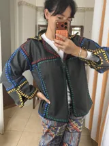 Chaqueta oversize de paño de lana en color beige, con costuras a la vista bordadas a mano en crochet con hilo multicolor en tonos rojos, azules, amarillos y verdes. Presenta cuello camisero y diseño amplio.