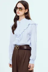 Camisa celeste de manga larga con sutiles rayas verticales, cuello alto mao fruncido y cierre frontal con botones. Presenta volados decorativos en los hombros y el frente, y puños elásticos fruncidos.