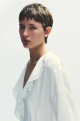 Vestido corto blanco de escote en V y manga larga con puños abotonados. Presenta volantes con bordados de encaje en el cuello, escote y falda. Cierre lateral con cremallera oculta.