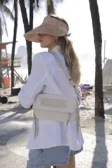 Cartera bandolera de cuero color beige, con solapa frontal decorada con detalles de costura y flecos colgantes en los laterales. Cuenta con correa ajustable de cuero con tachas.