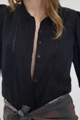 Camisa de lino color beige, de silueta romántica con escote a la base y mini volado. Cuenta con mangas 3/4 abullonadas con frunces y terminación en elástico. Posee detalles de puntillas negras en contraste en el frente y cierre frontal con botones ocultos bajo cartera.