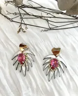 Aros colgantes con diseño de hoja plateada texturizada, coronados por un detalle en forma de corazón dorado. En el centro, presentan una piedra de cristal en forma de gota color fucsia, rodeada por pequeños cristales brillantes.