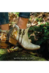 Bota corta de cuero color blanco y crema, con diseño pintado a mano en tonos dorados y marrones en la parte trasera. Cuenta con taco bloque de madera, puntera afinada y doble cierre lateral con tiradores metálicos personalizados.