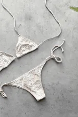 Conjunto de bikini con corpiño triangular de atar con apliques de argollitas y bombacha vedetina de atar con apliques de argollitas. El tejido presenta un estampado tie-dye o marmolado en tonos lila/azul y naranja/beige.