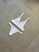 Conjunto de ropa interior compuesto por un bralette sin taza y una bombacha tipo tanga, confeccionados en algodón y lycra. El bralette presenta una banda elástica personalizada con el nombre de la marca y breteles anchos, mientras que la tanga cuenta con una banda elástica a juego.