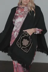 Ruana de tejido de punto color negro, con diseño abierto, flecos en el ruedo y bolsillos frontales destacados con bordados artesanales en tonos contrastantes.
