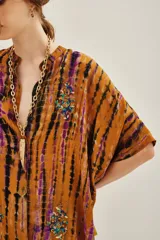 Camisa de manga corta con cuello mao y calce amplio, confeccionada en viscosa con estampado tie-dye en tonos ocre, negro y violeta. Presenta detalles bordados con mostacillas, lentejuelas, canutillos e hilos de lurex, y cierre frontal con botones a tono.