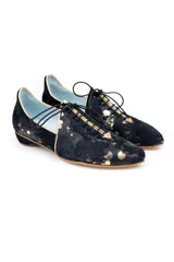 Zapato tipo blucher de pelo de pony en color negro con salpicaduras doradas. Presenta cierre de cordones, ribetes dorados a contraste y una silueta geométrica con aberturas laterales. Cuenta con un tacón bajo forrado en el mismo material.