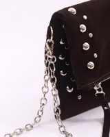 Bandolera de cuero gamuzado color marrón oscuro, con diseño rectangular y solapa frontal decorada con tachas metálicas plateadas. Incluye una cadena metálica plateada larga para usar al hombro o cruzada, y una cadena más corta para llevar como sobre.