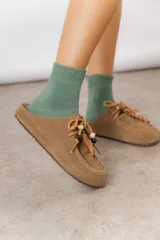 Zueco de cuero gamuzado color tostado, con cordones decorativos adornados con cuentas de madera y piezas envueltas en cuero. Diseño tipo mocasín con costuras visibles en la parte superior y abierto en la parte trasera.