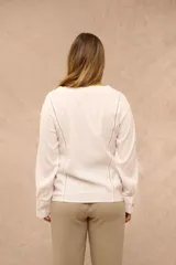 Blusa blanca de manga larga con escote en V y cierre frontal de botones. Presenta detalles de costuras caladas a lo largo del frente y en las mangas.