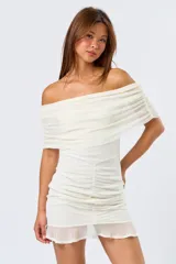 Vestido corto de mesh color blanco, con diseño off-shoulder y escote con volados. Presenta un ajuste drapeado y fruncido en la parte central de la falda, finalizando con un ruedo con volado sutil.