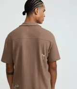 Camisa de manga corta, color marrón topo, con cuello camisero y corte amplio (boxy). Presenta bordados minimalistas en blanco en el frente, simulando un paisaje.