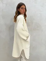 Sacón de punto color crudo con ribetes marrones, de corte largo y holgado, con cuello redondo, mangas largas y dos bolsillos frontales.