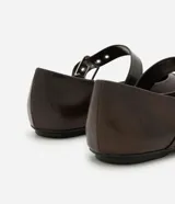 Zapatilla tipo bailarina color marrón oscuro, confeccionada en poliuretano con acabado mate. Presenta punta redondeada, suela plana y dos tiras ajustables con hebillas metálicas plateadas y ojales decorativos sobre el empeine.