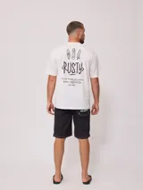 Remera blanca de algodón con cuello redondo y manga corta. Presenta un pequeño estampado frontal con el logo de Rusty y un diseño de tabla de surf.