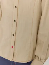 Camisa de denim con silueta levemente entallada, cuello bebé y mangas largas con sutil frunce en los hombros. Presenta costuras curvas decorativas con pespuntes en contraste y cierre frontal con botones.