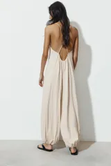 Mono largo de corte holgado estilo bombacho, color beige, con escote redondo, tirantes finos cruzados en la espalda con detalle de abalorios de madera y bajos ajustados con puños elásticos.