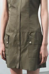 Vestido corto de estilo utilitario en color verde militar, confeccionado en gabardina. Presenta cuello redondo, cierre frontal con cremallera oculta, mangas cortas con detalle de trabilla y botón, y bolsillos de plastrón en el frente.