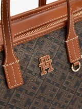 Cartera tote Tommy Hilfiger de acabado texturizado con estampado integral del monograma TH en tonos marrón y negro. Cuenta con dos asas superiores de cuero sintético tostado, bandolera extraíble, cierre de cremallera y herrajes metálicos dorados. Incluye una insignia del monograma en el frente y un detalle de anilla en D.