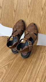 Sandalias de cuero negro con diseño de tiras trenzadas artesanales en la parte delantera, plantilla de cuero y tira ajustable al tobillo con hebilla metálica.