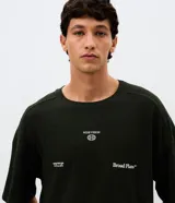 Remera de corte oversized color verde oscuro, con cuello redondo y mangas cortas. Presenta un diseño estampado en la espalda con tipografía en blanco y un pequeño logo gráfico en la parte inferior.