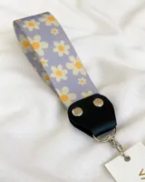Llavero de tela con estampado de flores blancas y amarillas sobre fondo lila. Tiene un conector de cuero sintético negro y un gancho metálico plateado.