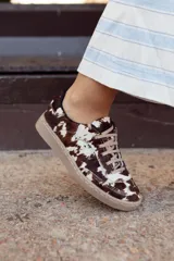 Sneaker de diseño artesanal confeccionada en cuero con pelo natural con estampado de manchas tipo vaca en tonos marrón y blanco. Presenta cordones al tono y suela de goma color caramelo.