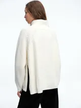Sweater de tejido de punto acanalado en color blanco, con cuello alto y cierre metálico diagonal en el hombro. Presenta mangas largas con diseño raglán y corte holgado.