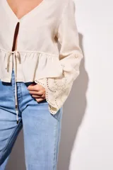 Blusa color beige claro, con escote en V profundo y cierre frontal mediante lazo ajustable a la altura de la cintura. Presenta un peplum con volado y mangas largas con puños anchos con bordados calados.