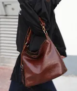 Cartera tipo hobo de cuero marrón tabaco con textura grabada, cierre principal superior y correa de hombro ajustable con herrajes metálicos.