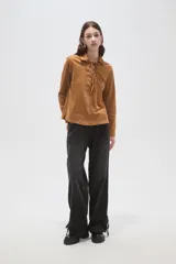 Blusa de gamuza sintética color camel, con cuello camisero y cierre frontal mediante cordones entrelazados. Presenta mangas largas y un corte holgado.