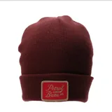 Gorro tipo beanie de tejido de punto en color bordó, con vuelta y parche rectangular frontal en color rojo con bordado en tono beige.