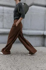 Pantalón de vestir marrón oscuro, tiro alto, con pernera ancha y pinzas frontales. Presenta un bolsillo con tapeta en la parte superior delantera.