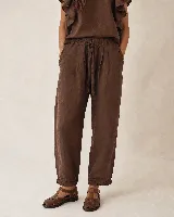 Pantalón de lino marrón con silueta relajada y amplia, cintura elastizada con lazo ajustable, bolsillos laterales y detalles de pliegues en los laterales y el ruedo.