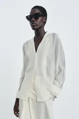 Camisa de mujer fluida de ZARA WOMAN COLLECTION, confeccionada en hilatura con mezcla de lyocell. Presenta escote en pico, manga larga con puños marcados y bajo asimétrico, con cierre frontal de botones ocultos.