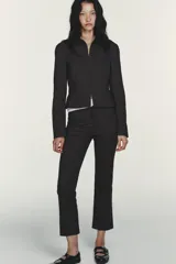 Pantalón entallado de tiro alto color negro, con cintura con trabillas y bolsillos de vivo con cremallera en el delantero. El bajo tiene un acabado en línea evasé. Cierre frontal con cremallera, botón interior y gancho metálico.