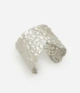 Pulsera tipo brazalete rígido de metal plateado con acabado martillado y textura irregular. Presenta un diseño asimétrico con abertura frontal.