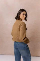 Chaqueta de gamuza sintética color tostado, con cuello mao, cierre frontal con cremallera y bolsillos frontales con solapa. Presenta detalles de broches metálicos y puños ajustables.