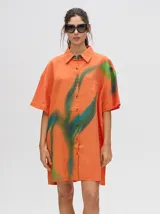 Camisa tipo vestido de corte oversize, confeccionada en viscosa. Presenta un diseño estampado con trazos abstractos en tonos verdes sobre una base naranja, cuello clásico, cierre frontal con botones y mangas cortas.