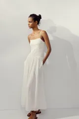 Vestido midi de color blanco con escote recto y tirantes finos ajustables. Presenta un cuerpo con tejido elástico tipo nido de abeja y detalles de flores bordadas en la parte superior. Cuenta con bolsillos laterales ocultos y forro interior.