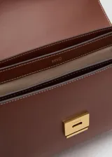 Bolso de hombro tipo bandolera en color marrón, con diseño estructurado de solapa, cierre metálico a presión y costuras decorativas en contraste. Incluye correa larga ajustable.