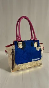 Cartera de cuero estilo tote de la marca Carolina De Cunto, confeccionada con una combinación de cueros en tonos azul, fucsia, beige y dorado. Cuenta con asas de mano fucsias, detalles de pelo sintético en el frente, bolsillos externos con cierre y herrajes metálicos.