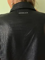 Camisa negra de cuero sintético con textura de cocodrilo, corte recto, cuello clásico, cierre frontal con botones metálicos y bolsillos con solapa en el pecho.
