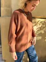 Sweater de punto color terracota, con diseño holgado, cuello redondo y aberturas laterales en el ruedo. Presenta puños acanalados y una etiqueta decorativa en la parte inferior.