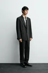Pantalón de vestir negro con pliegues frontales, corte regular fit, bolsillos laterales y traseros de doble vivo con botón. Cierre frontal cruzado con cremallera y gancho metálico.