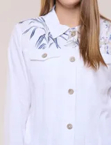 Campera liviana de lino color blanco, con estilo denim y silueta semi entallada. Presenta un canesú superior estampado con motivos florales en tonos azules y tostados, cuello camisero, abotonadura frontal con botones rústicos y bolsillos con solapa en el pecho.