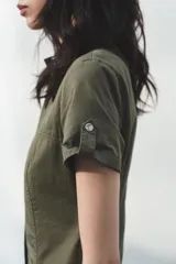 Vestido corto de estilo utilitario en color verde militar, confeccionado en gabardina. Presenta cuello redondo, cierre frontal con cremallera oculta, mangas cortas con detalle de trabilla y botón, y bolsillos de plastrón en el frente.