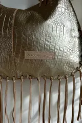 Cartera de hombro color dorado con textura símil cocodrilo y flecos colgantes en los bordes y las asas, de la marca Carolina de Cupto.