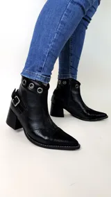 Bota corta de cuero color negro, con diseño estilo tejano, punta fina y taco cuadrado. Cuenta con detalles de ojales metálicos en el borde superior, una hebilla lateral ajustable y cierre interno.
